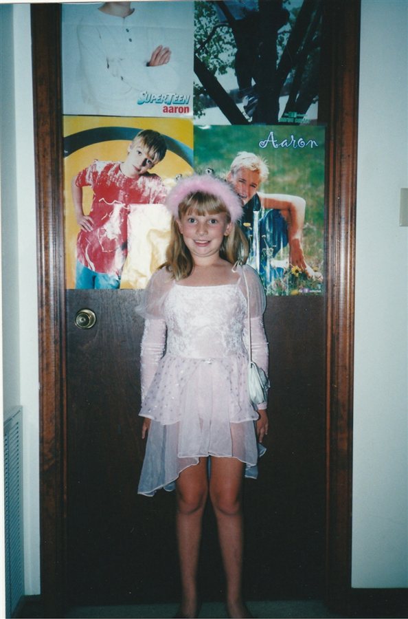 Hannah as a kid in a dress-up gown, posing in front of her Aaron Carter poster door